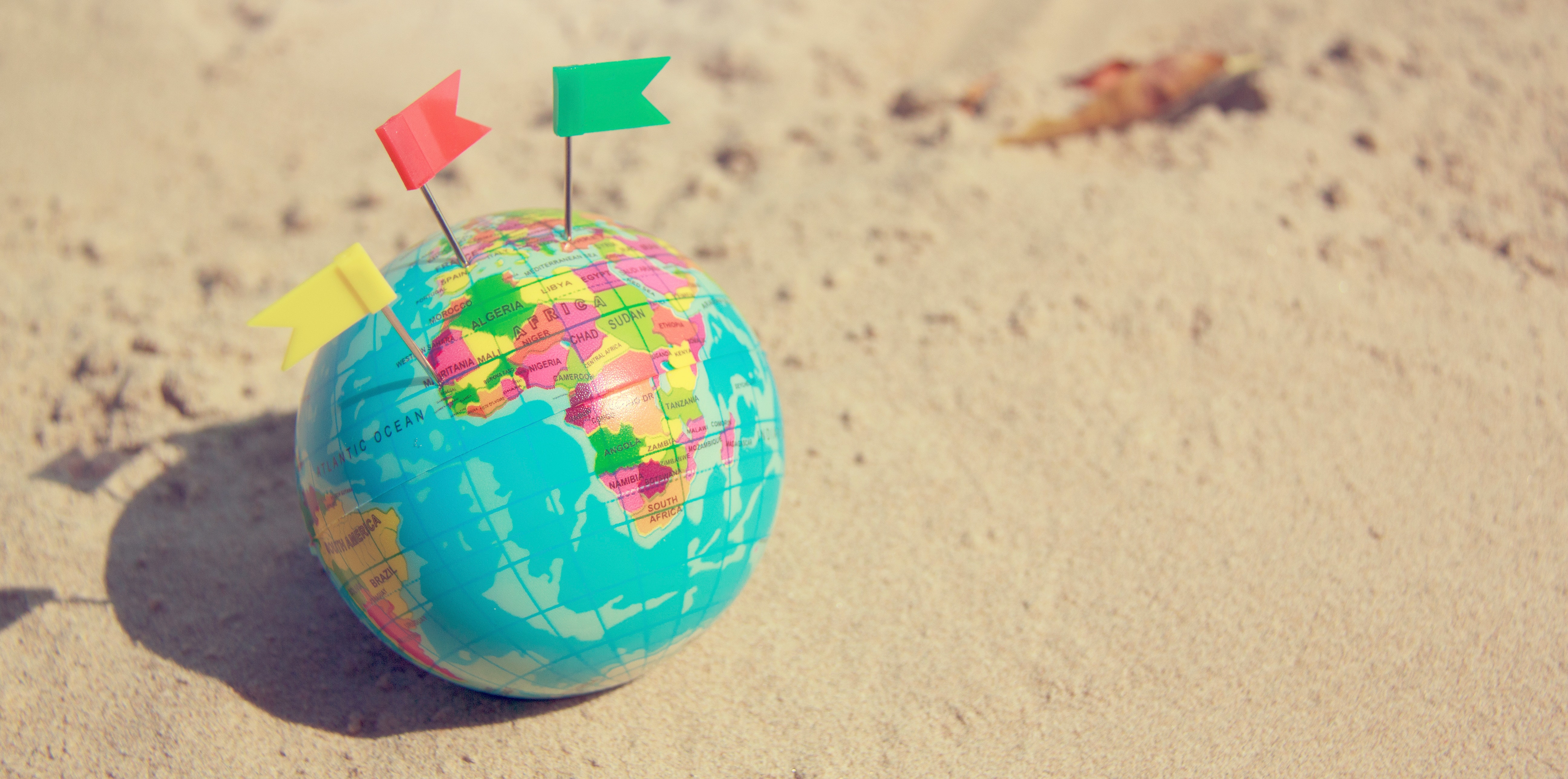 Small globe with three colorful flag pins sitting on the sand.